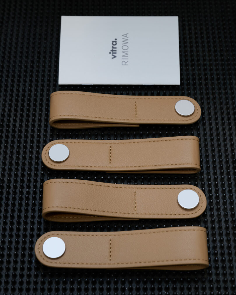 Four tan leather straps with silver snaps are laid out in a row below a white card labeled 