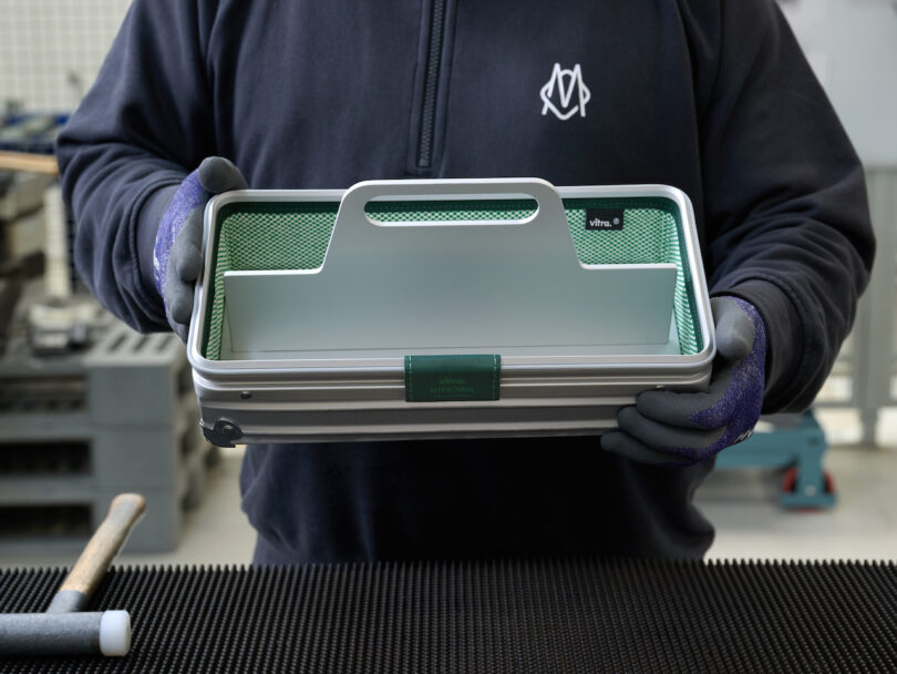 A person wearing gloves and a dark jacket holds a rectangular metal and mesh toolbox with a handle and green accents. A hammer and black mat are visible on the work surface