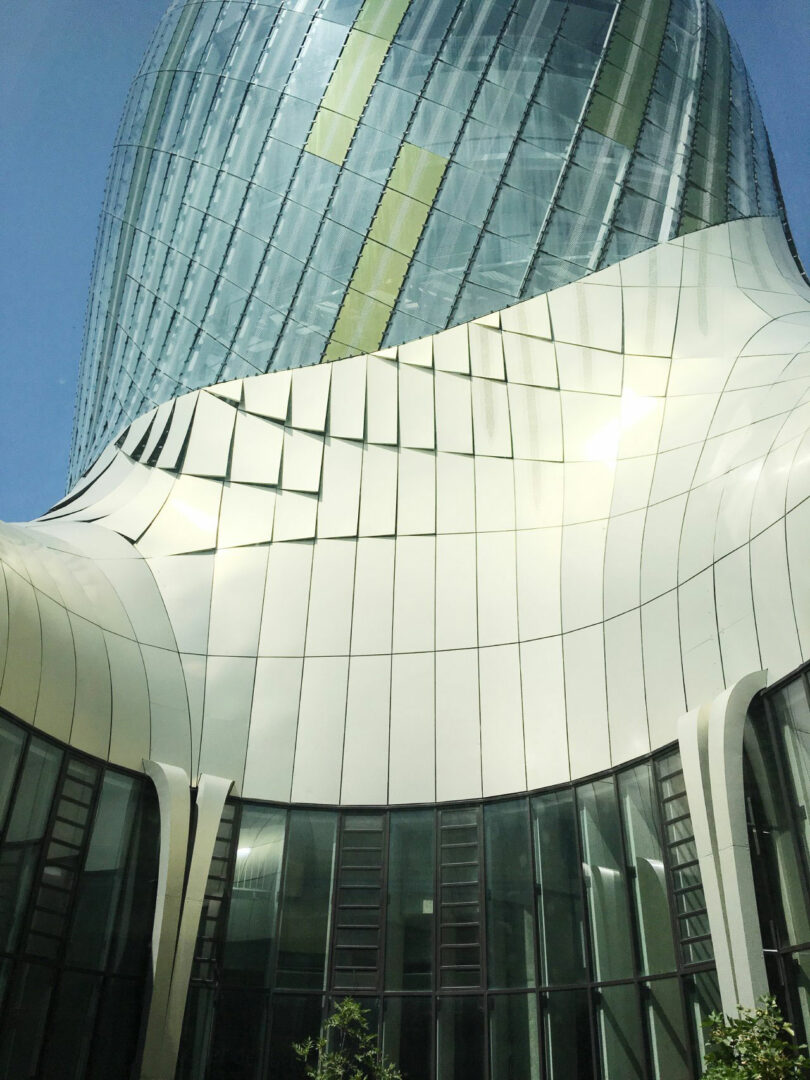 A modern building with a curved, reflective glass and metal facade, featuring vertical windows and contemporary architectural design by Tina Ramchandani against a blue sky.