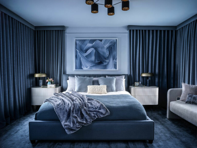 A modern bedroom by Tina Ramchandani with blue walls, blue bedding, blue curtains, abstract wall art, two nightstands with lamps, and a gray sofa on the right.