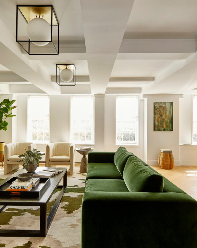 A modern living room designed by Tina Ramchandani features a green velvet sofa, light armchairs, a coffee table with books and plants, large windows, abstract art on the wall, and geometric ceiling lights.