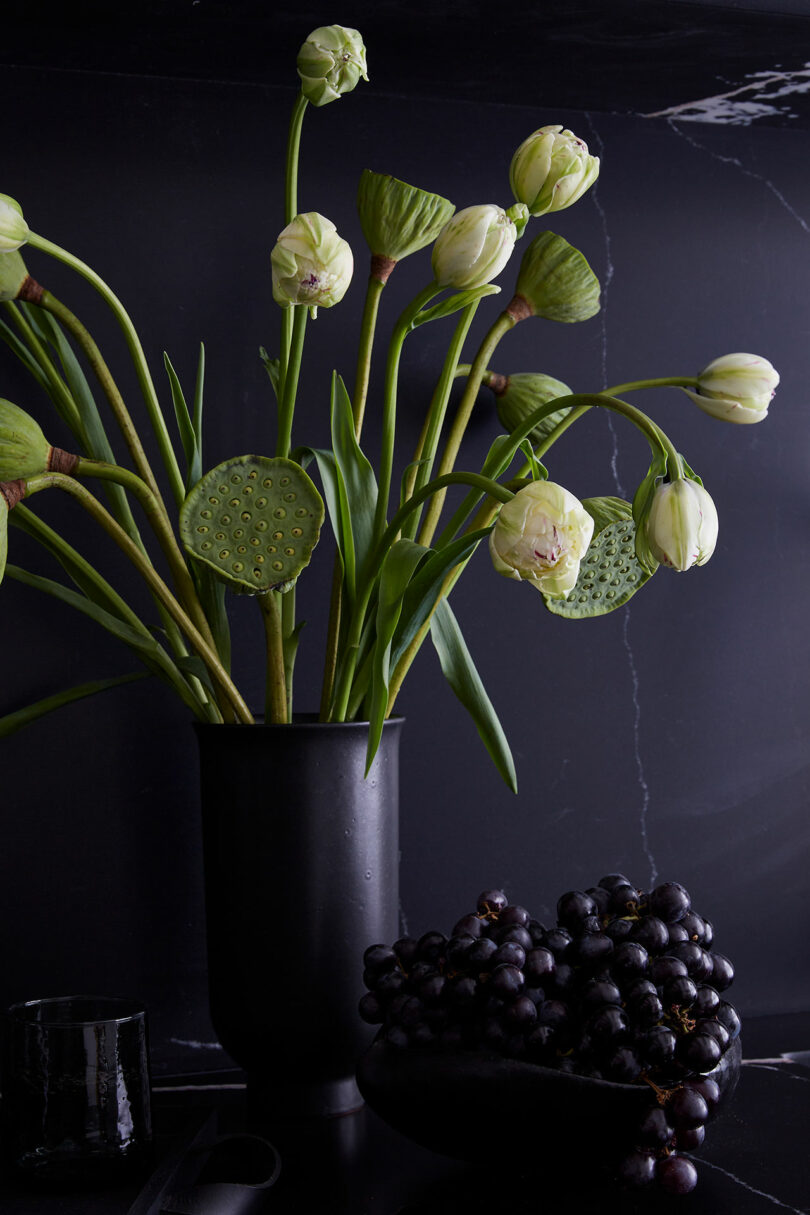 A black vase holds white tulips and lotus pods next to a bowl of black grapes, set against a dark background in a style reminiscent of Tina Ramchandani’s sophisticated designs.