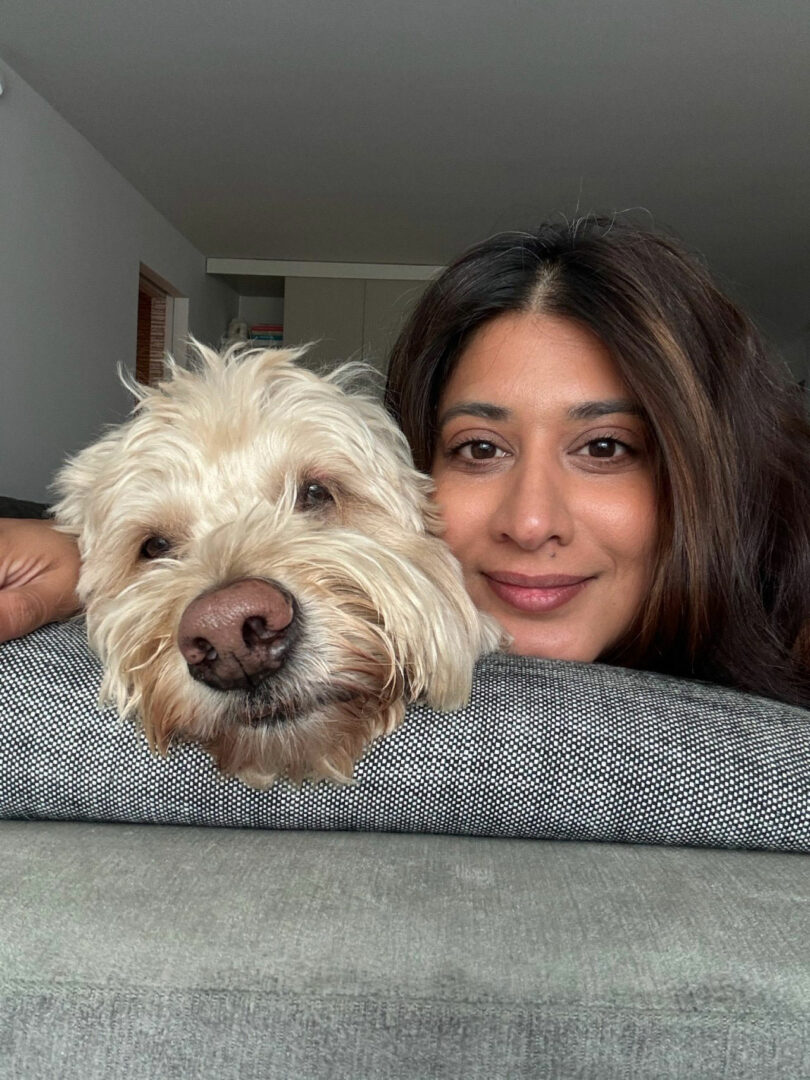 Tina Ramchandani and a fluffy light-colored dog rest their heads together on a gray couch, both looking at the camera.