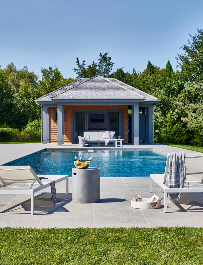 A backyard pool with two lounge chairs, a side table, and towels in the foreground; inspired by Tina Ramchandani, this serene retreat features a pool house at the far end, surrounded by greenery under a clear sky.