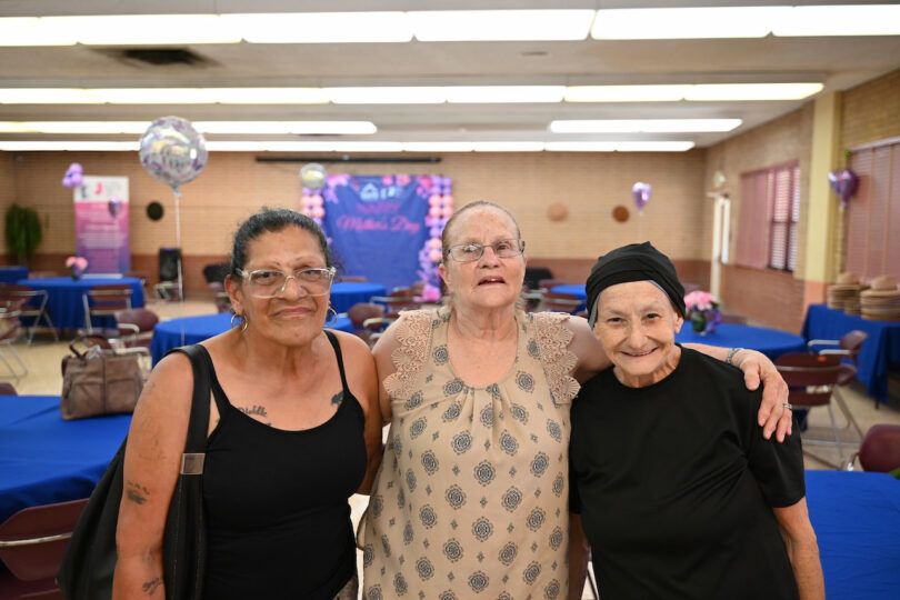 Three elderly women pose together and smile in a decorated event hall with blue tablecloths, balloons, and pink-themed decorations in the background