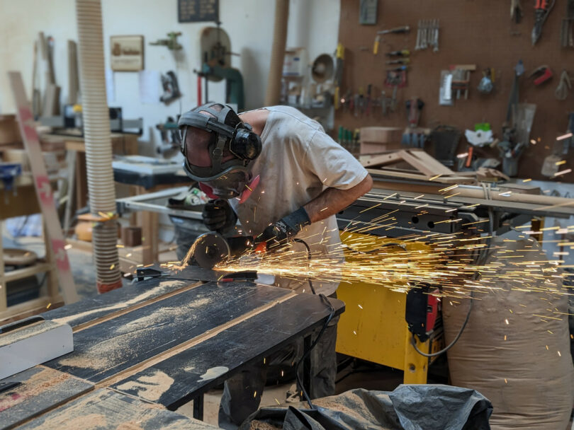 A person wearing safety gear uses a power tool to cut metal in a workshop, producing bright sparks that fly across the room