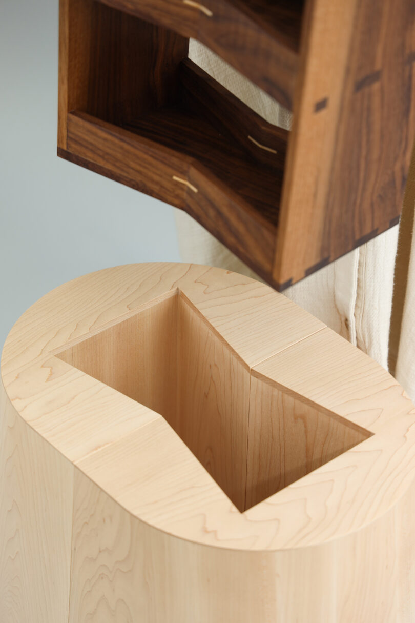 A person places a small, rectangular wooden box with shelves into a matching slot on a light-colored wooden base
