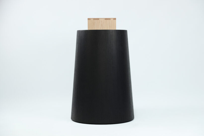 Black, asymmetrical wooden stool with a flat, light wood inlay on the top, set against a plain white background