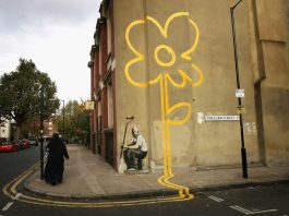 A woman walks past a some a painting of a yellow flower on a wall on a street.