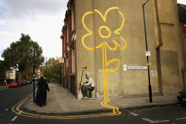 Banksy-Mural-at-the-Center-of-Legal-Battle.jpg A woman walks past a some a painting of a yellow flower on a wall on a street.