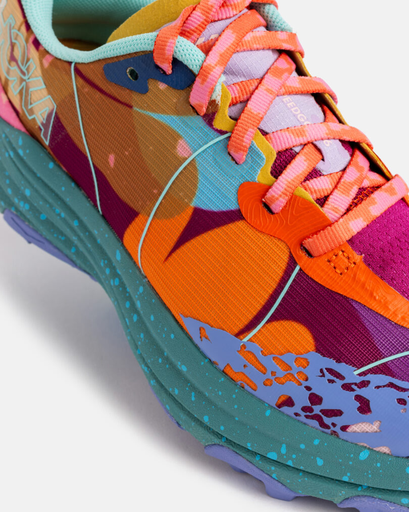 Close-up of a colorful running shoe with a multicolored upper, speckled teal sole, orange accents, and coral pink laces. The brand name 
