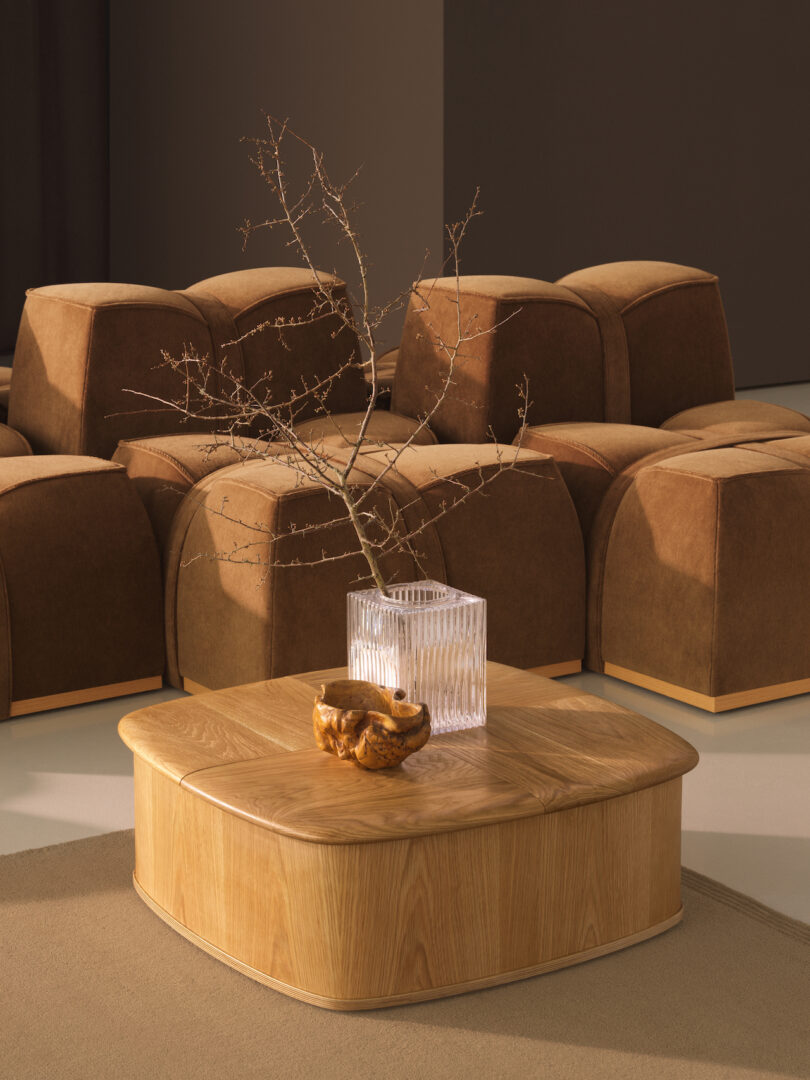 A wooden coffee table with a glass vase holding bare branches and a carved wooden bowl, set in front of brown upholstered ottomans