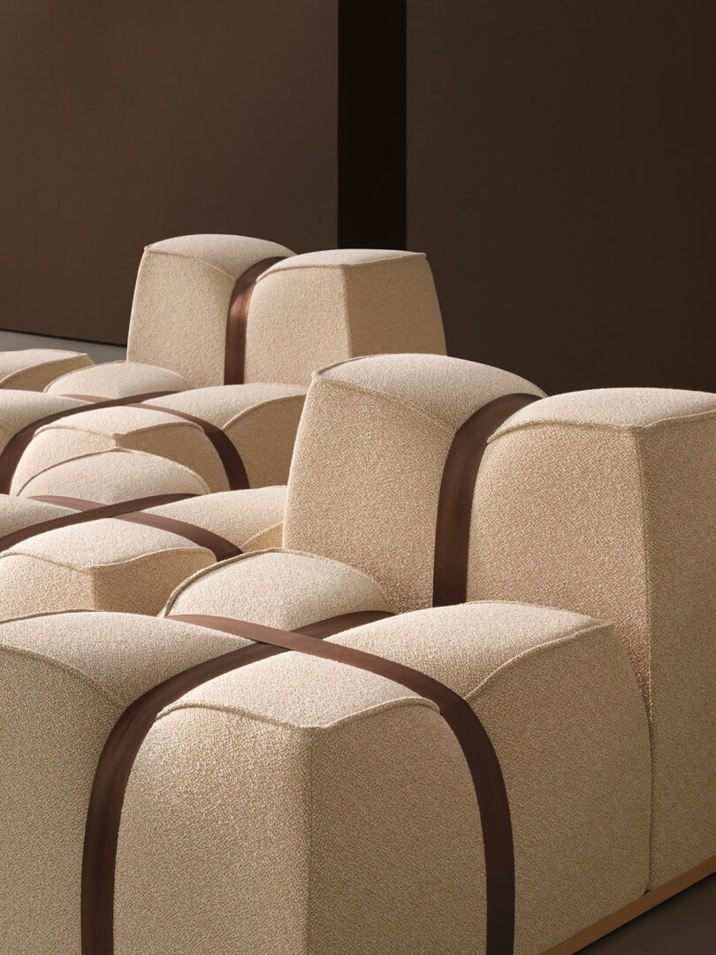 Beige, block-shaped upholstered seats by Mizetto are arranged in a grid pattern, with dark brown straps intersecting over the tops, set against a brown background.