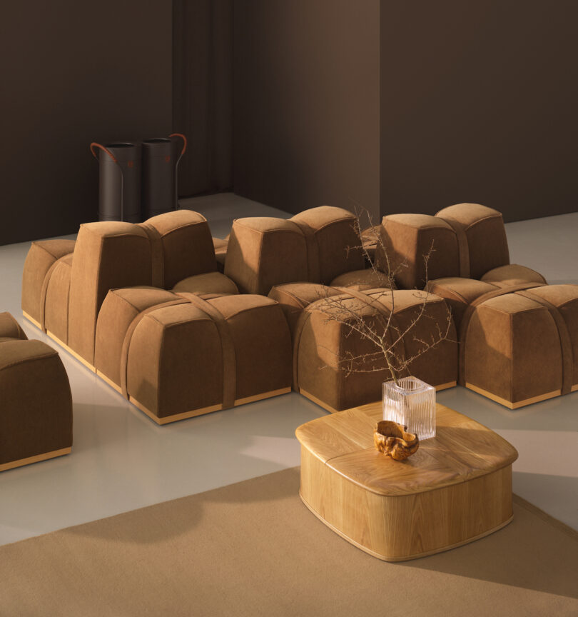 A minimalist room with brown modular seating, a low wooden table with a glass vase and dried branch, and two black bins in the corner against a brown wall