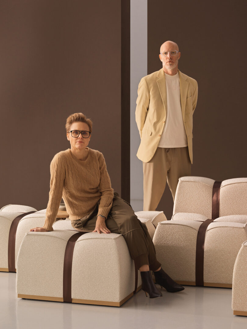 Two people in neutral-toned outfits pose among cream-colored, ribbon-wrapped seating blocks against a brown backdrop. The person sitting wears glasses and a sweater; the person standing wears a blazer