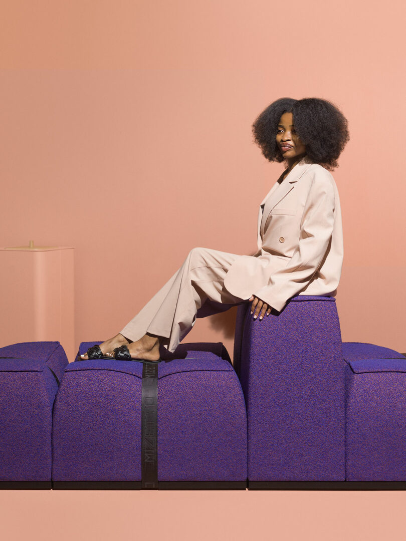 A woman in a light beige suit sits sideways on a blocky, purple Mizetto modular sofa against a peach-colored background.