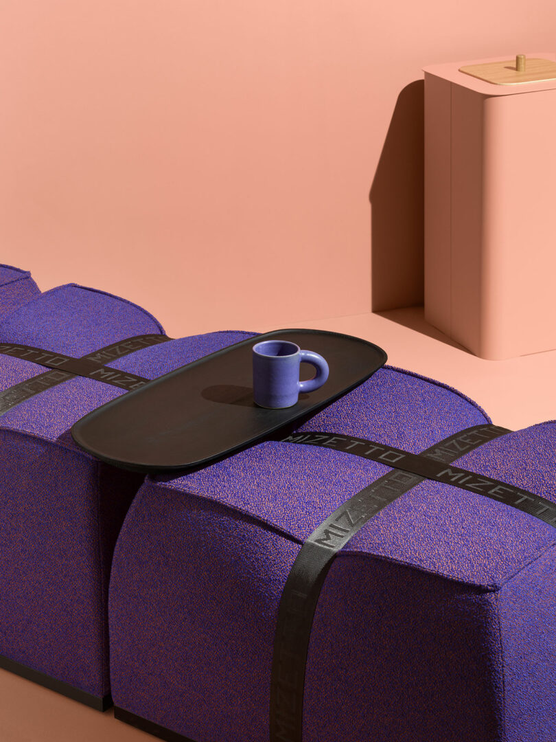 A Mizetto purple mug on a black tray rests atop two textured purple ottomans with black straps, set against a pink background with a pink side table in the corner.