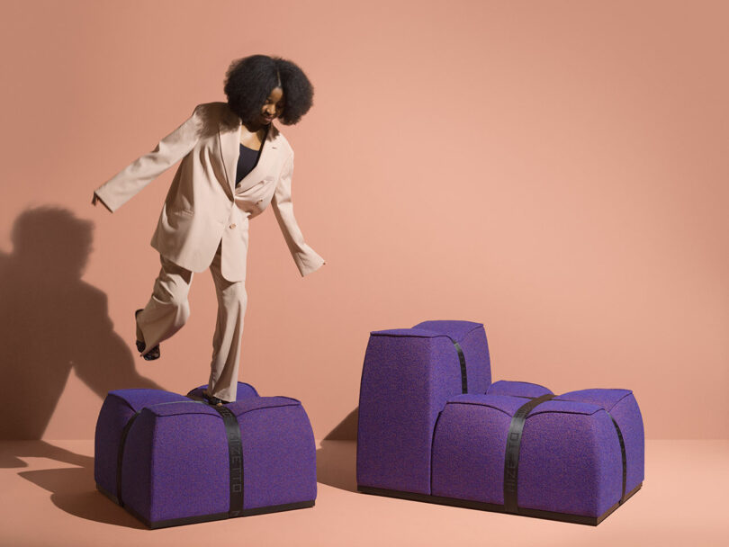 A person in a beige suit balances on large, purple, cushion-like Mizetto blocks against a plain peach background.