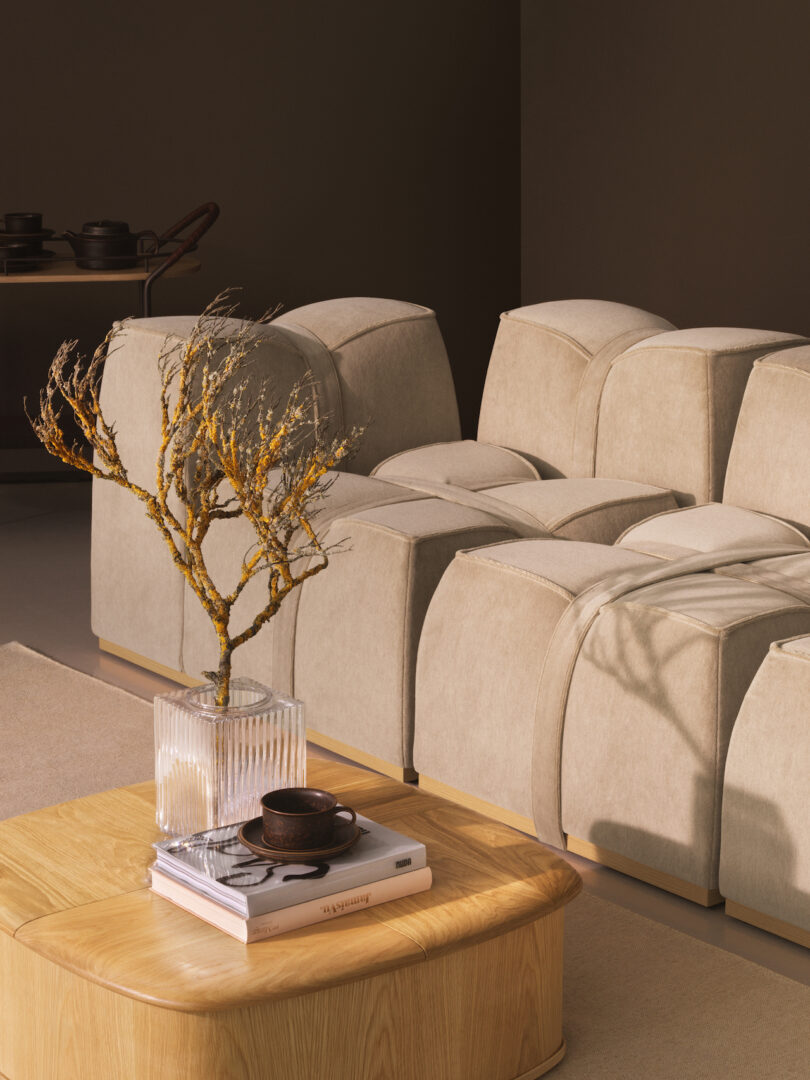 A modern living room with a beige modular sofa, a wooden coffee table holding a glass vase with dried branches, a book, and a dark ceramic cup and saucer