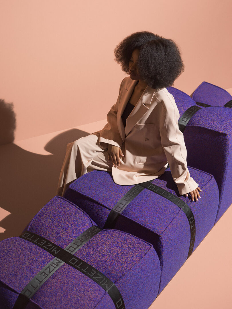 A woman in a beige suit sits on large, Mizetto purple block-shaped seats with black straps against a peach-colored background.