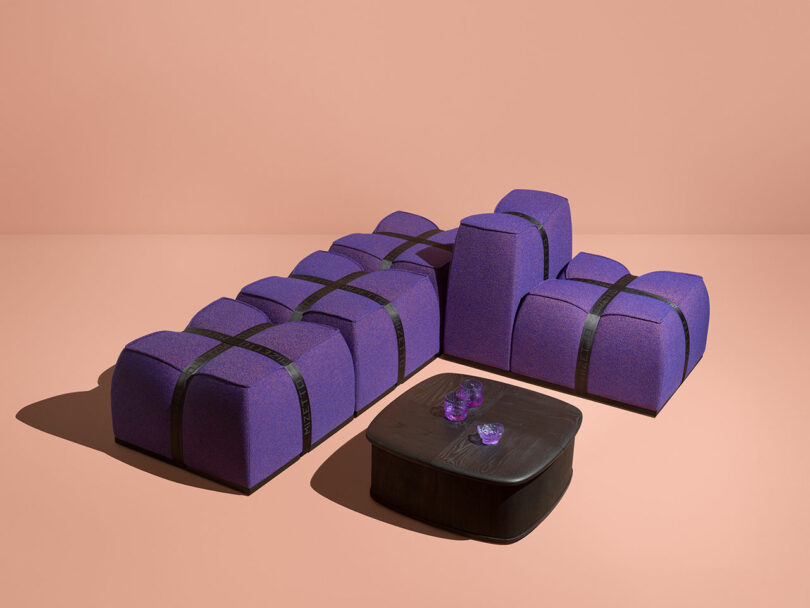 A modular Mizetto purple sofa with black straps is arranged in an L-shape next to a low black coffee table topped with three amethyst crystals, set against a plain peach background.