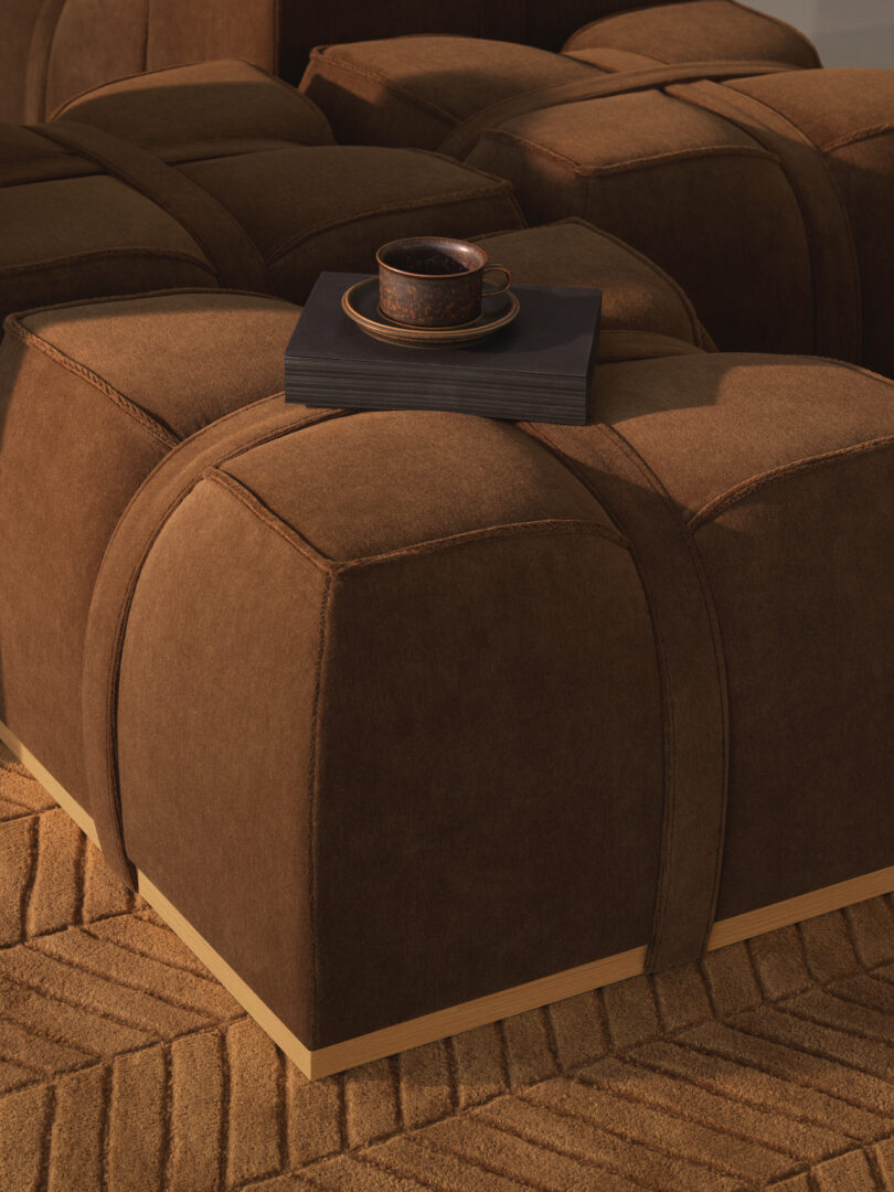 A brown, modular upholstered ottoman with a matte black tray holding a cup and saucer, placed on a textured tan carpet.