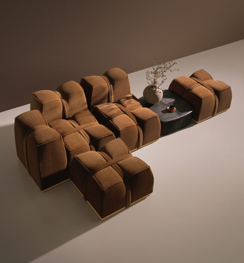 A modular brown sectional sofa with plush cushions is arranged around a black coffee table holding a beige vase with branches and a small round item, set against a minimalist brown background