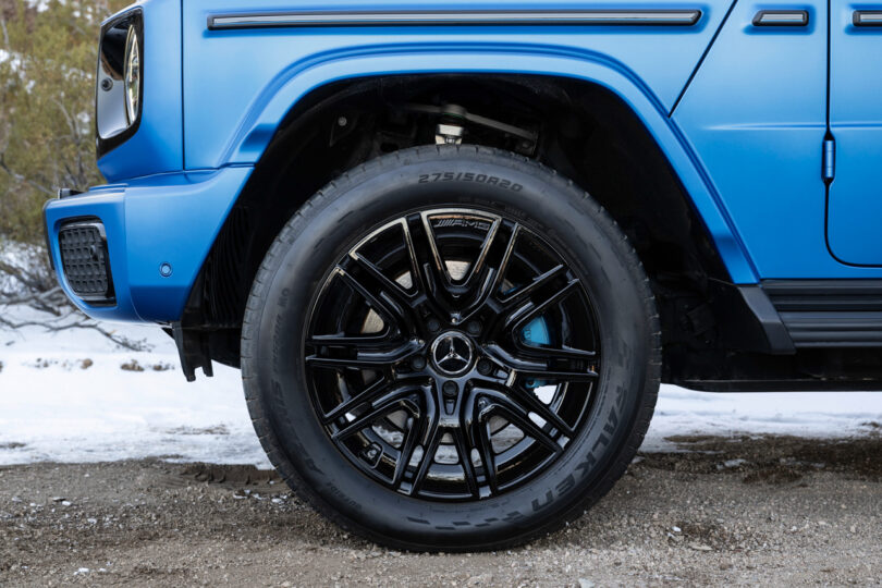 Close-up of the front left wheel of a blue Mercedes-Benz G580 SUV, featuring a black multi-spoke rim and Falken tire, with snowy ground and vegetation in the background.