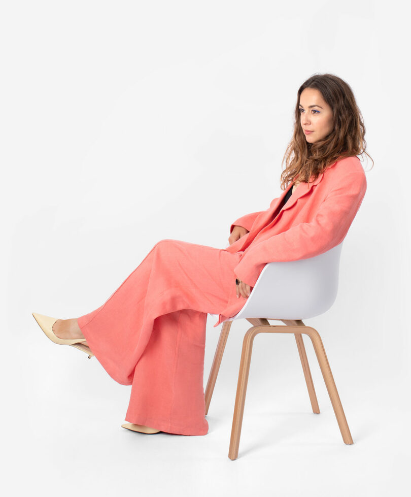 Woman in a coral-colored suit sits sideways in a modern white chair with wooden legs, looking ahead, beneath the soft glow of the Light Bean by Katerina Kopytina, against a plain white background.