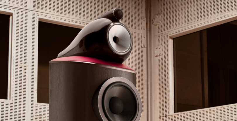 A close-up of a high-end Bowers & Wilkins speaker system with a rounded design, shown against a backdrop of soundproof acoustic panels and large windows.