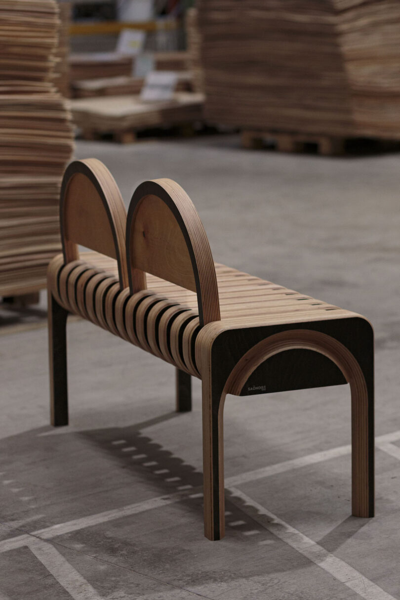 A modern wooden bench with two arched backrests, made of layered plywood, sits on a concrete floor in a warehouse with stacks of wood in the background