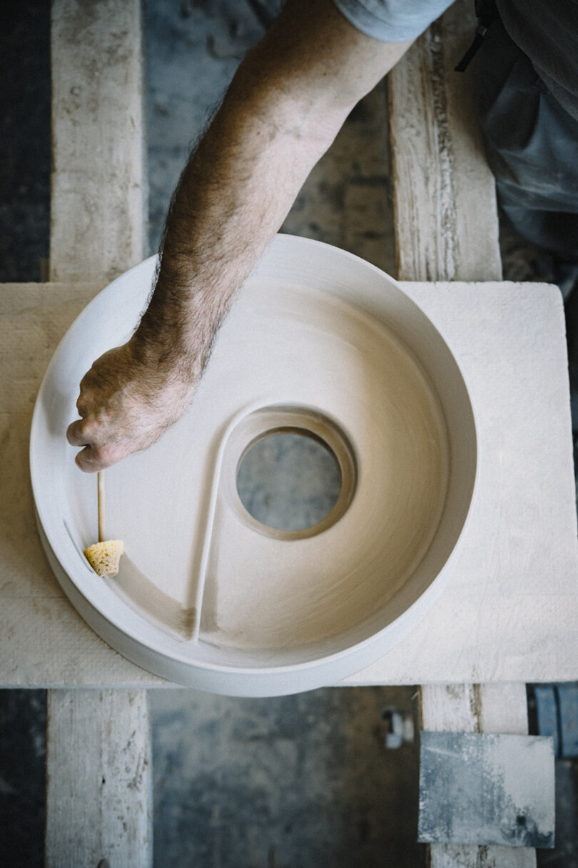A person’s arm is smoothing the surface of a round ceramic sink with a sponge on a wooden workbench.