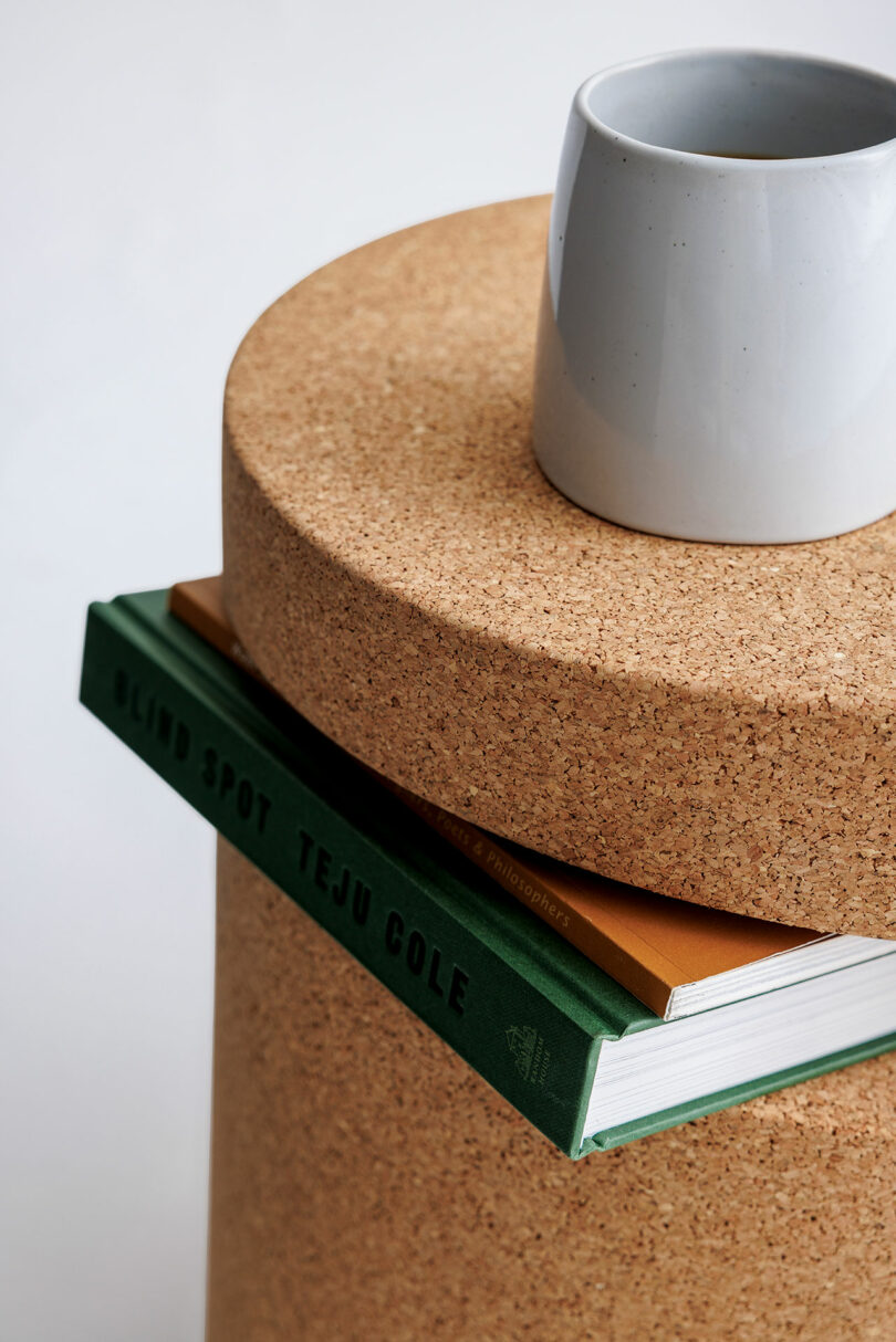 A grey ceramic cup sits on a round cork table, with a green book titled 