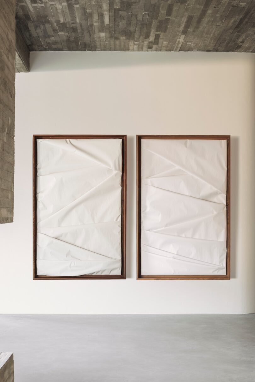 Two large wooden frames containing crumpled white material hang side by side on a plain wall in a minimalist room with a concrete ceiling.