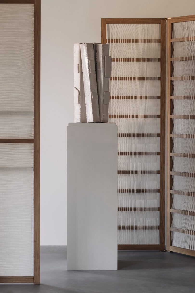 A white abstract sculpture sits on a tall pedestal, flanked by wooden room dividers with semi-transparent, textured panels in a minimalist interior setting.