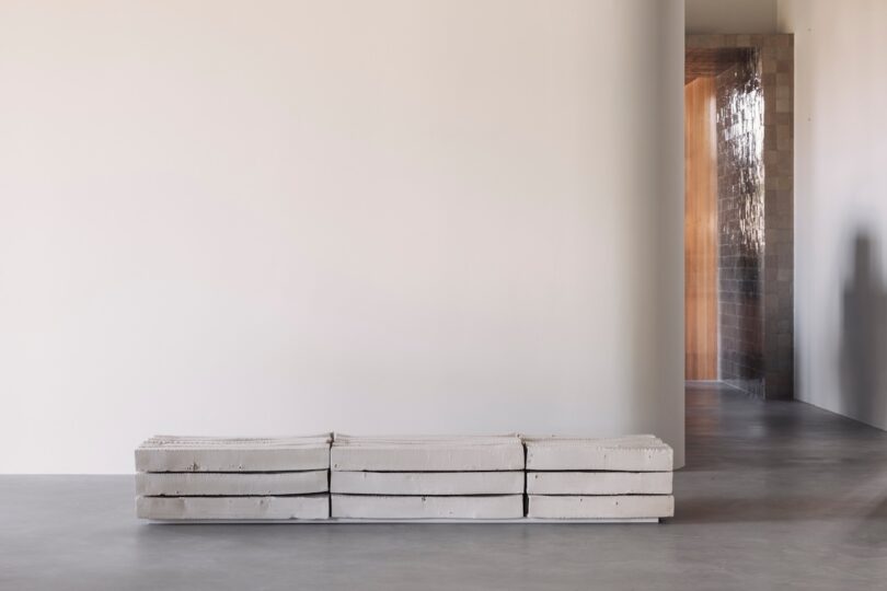 A minimalist interior with a concrete bench against a plain white wall, and a hallway with textured walls and soft light in the background.
