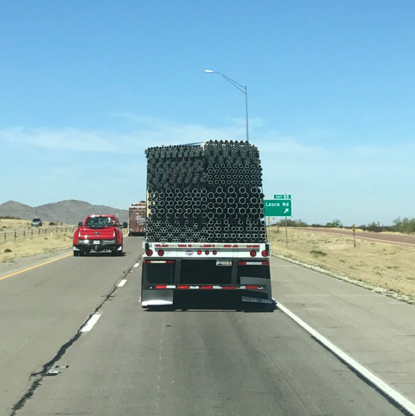 A truck carrying stacked black pipes drives on a highway; a red pickup follows behind. A green road sign for