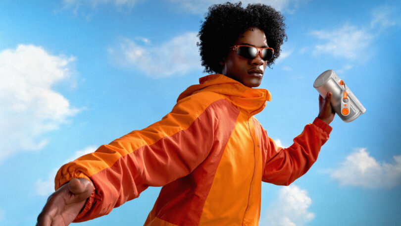 A person wearing an orange jacket and sunglasses holds an XGIMI portable speaker against a blue sky with clouds.