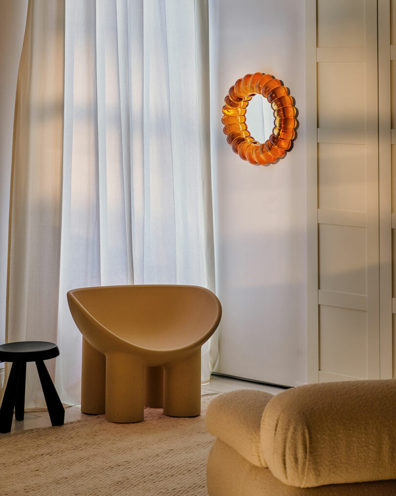 A modern room with a sculptural beige chair, a small black stool, white curtains, a round orange wall mirror, and a textured beige sofa