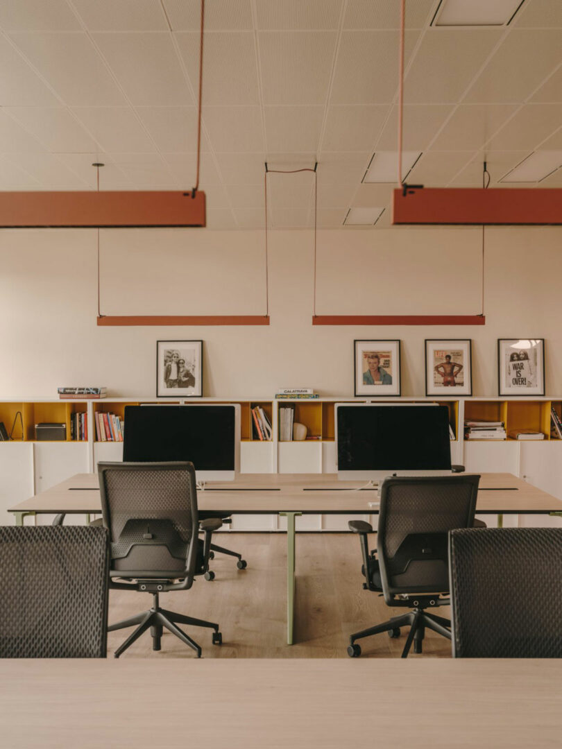 Modern office space with empty desks, black office chairs, large monitors, shelving with books, and framed photos on the back wall.