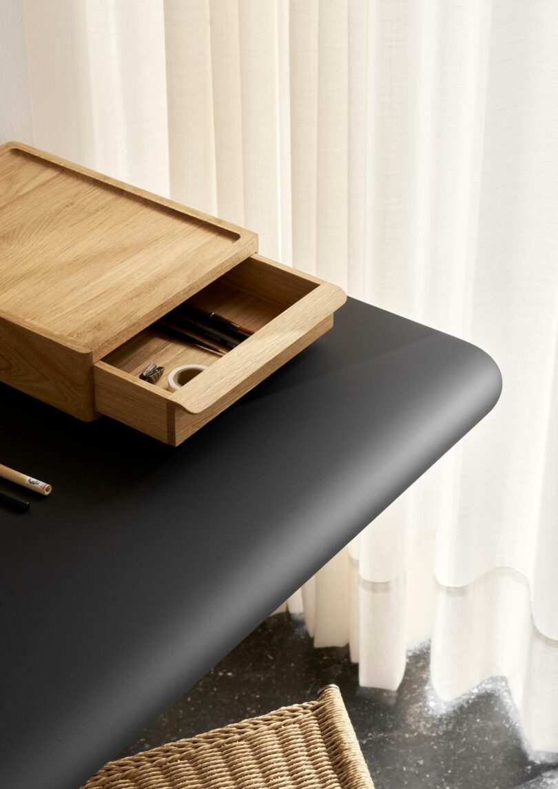 A black desk with a woven Carl Hansen & Son Anker Bak chair, a wooden organizer box with an open drawer containing pens and small items, next to sheer white curtains