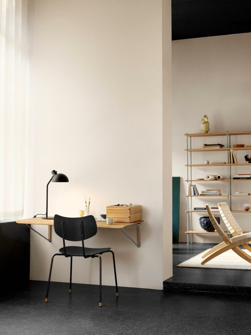 Minimalist home office setup featuring a Carl Hansen & Son Anker Bak wall-mounted wooden desk, black chair, desk lamp, and shelves with decorative items in a bright, modern room