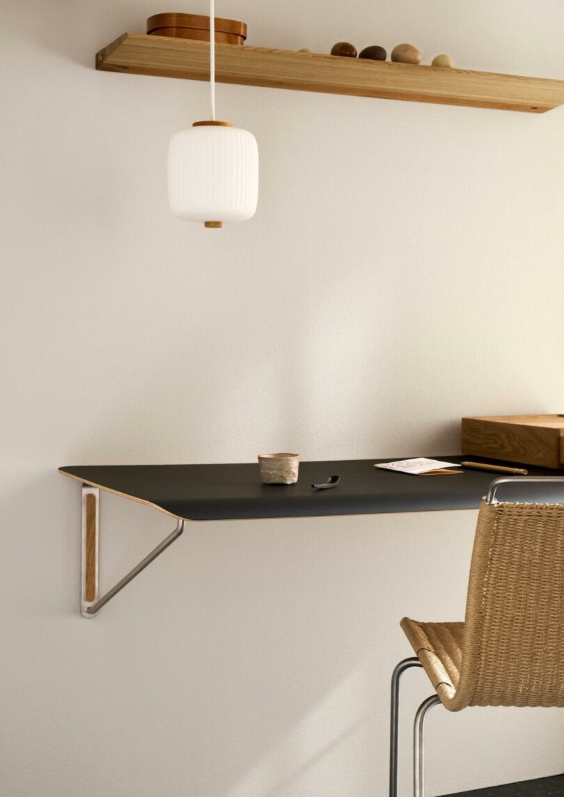 Minimalist workspace featuring a Carl Hansen & Son Anker Bak floating black desk, a woven chair, a hanging white lamp, a wooden shelf with decor accents, plus a cup and pen neatly arranged on the desk