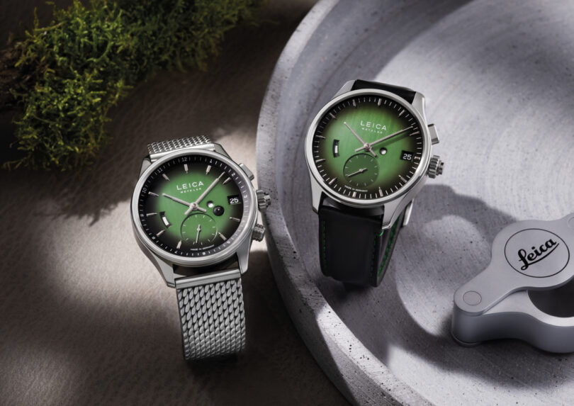 Two Leica wristwatches with green dials are displayed; one features a metal mesh band, the other a black leather strap. A Leica-branded item and moss add to the stylish, photography-inspired scene.