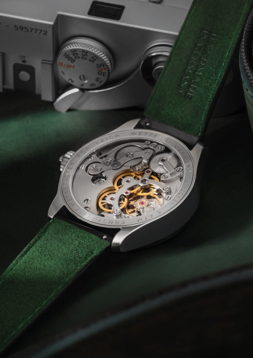 A mechanical wristwatch with a green leather strap displays its intricate movement, positioned next to a silver Leica camera on a green surface.