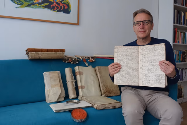 Art-Detective-Recovers-Documents-Stolen-From-Netherlands-Archives.avif.avif A man with glasses sits on a blue couch amongst old historic documents.