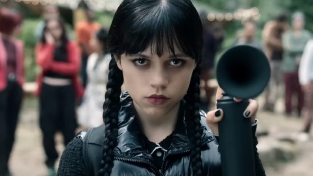 Dark-visions-and-monsters-abound-in-Wednesday-S2-trailer.jpg dark haired goth girl in pig tails dressed all in black holding up an emergency horn