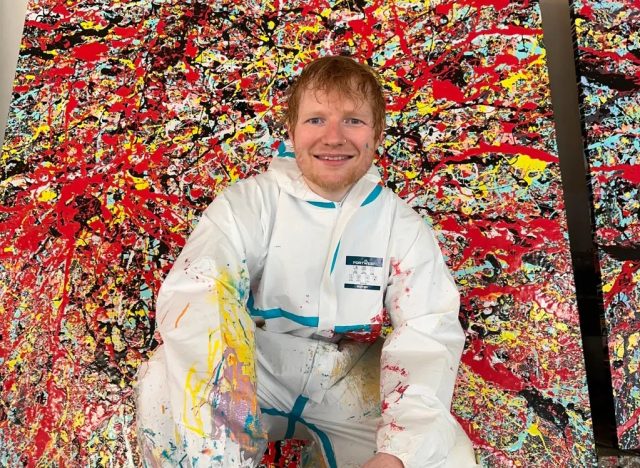 A painting of a man in coveralls kneeling in front of an abstract painting with splatters of red and blue paint.