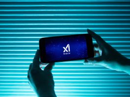 A person holds a telephone displaying the logo of Elon Musk s artificial intelligence grok in front of a background lit by a blue light.