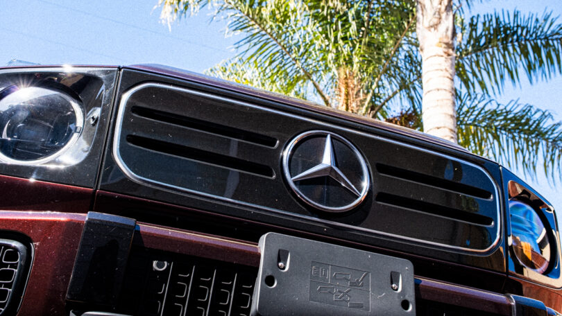 Close-up of the front grille and emblem of a Mercedes-Benz G580 SUV, with a palm tree and blue sky in the background.